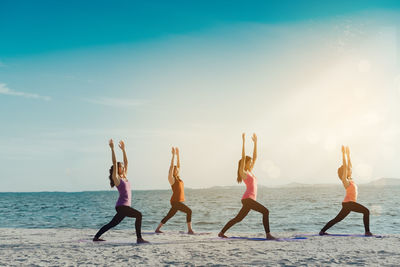 Female friends practicing yoga in warrior position on shore at beach during sunny day