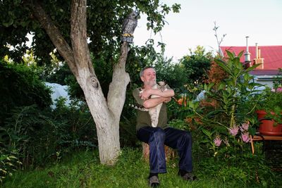 Senior man holding cat while sitting on tree stump in yard