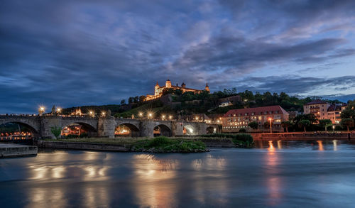 Illuminated bridge over river by buildings against sky at dusk