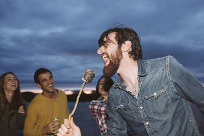 Smiling friends looking at man singing while holding roasted marshmallow on stick against cloudy sky