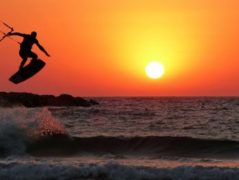 Silhouette man kitesurfing over sea against orange sky during sunset