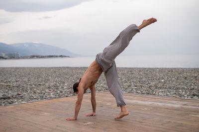 Shirtless caucasian man doing backflip on pebble beach. 