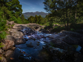 Stream flowing through rocks by river against sky