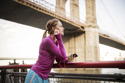 Woman listening music while standing against brooklyn bridge