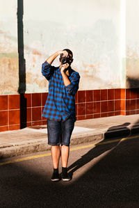Full body of male in casual outfit taking photo while standing on asphalt street under sunlight