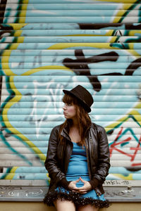 Woman wearing hat looking away against graffiti shutter in city