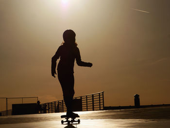 Silhouette woman skateboarding on footpath against sky during sunset