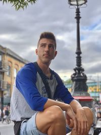Young man looking away while sitting in city against cloudy sky