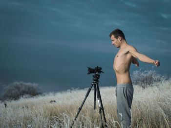 Side view of shirtless man with arms outstretched standing by camera on tripod in grassy field against sky