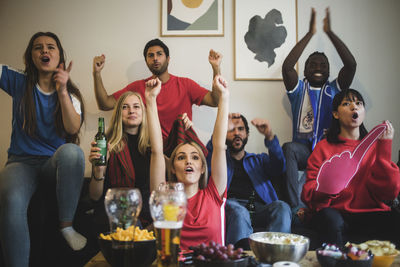 Happy friends with arms raised watching sport while sitting on sofa at home