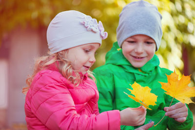 Smiling sibling wearing warm clothing holding leaf standing outdoors
