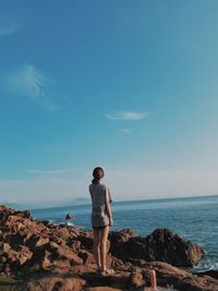 Rear view of young woman standing on rock by sea against sky