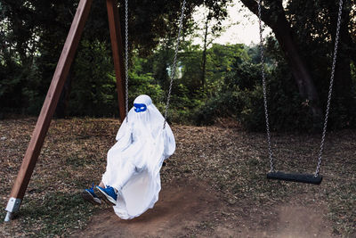 Unrecognizable kid in white ghost costume swinging and playing on playground during halloween holiday