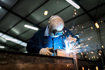 Unrecognizable male employee in protective gloves and helmet using welding machine while working in dark workshop