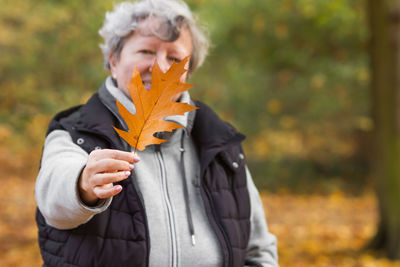 Happy senior woman covering her face with brown oak leaf in the autumn city park. nature background.