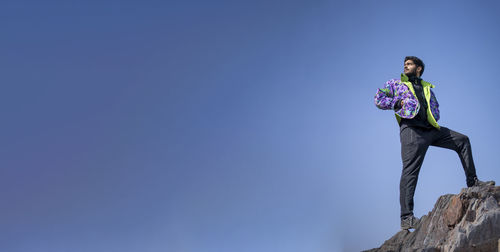 Low angle view of man standing on rock against clear blue sky