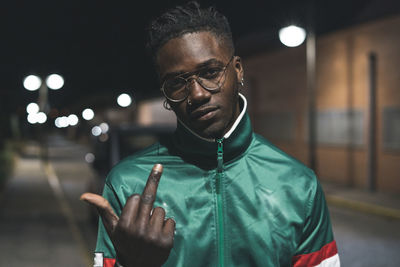 Portrait of young man showing middle finger while standing outdoors at night