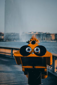 Close-up of coin-operated binoculars on pier against sky. fountain of geneva in the background.