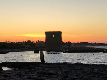 Silhouette building by sea against orange sky