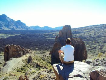 Hiker looking at mountain while sitting on rock against clear sky during sunny day