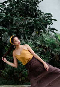 Young woman looking away while sitting on plant against trees