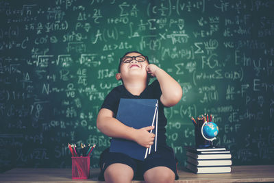 Cute boy holding books while sitting at table against blackboard