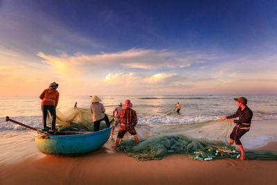 Fishermen fishing on shore at beach against sky during sunset