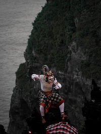 Rear view of man sitting on rock by sea against mountain