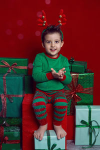 Baby boy in a green t shirt and with deer antlers sits with gifts on a red background