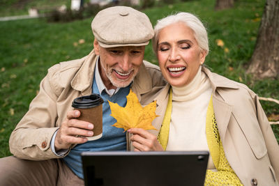 Smiling senior man holding autumn leaf and coffee cup talking on video call at park