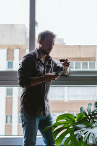 Young man using smart phone while standing against window