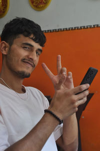 Closeup of a happy young guy looking at camera with showing peace sign while using mobile phone