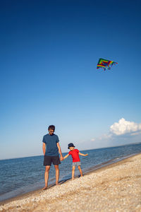 Father and son are standing on a sandy beach by the sea and launch a toy striped kite in the summer