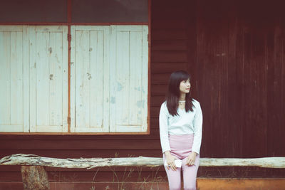 Young woman looking away while standing against wooden wall