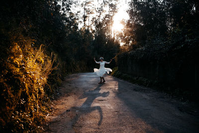 Full body of anonymous female wearing white dress walking on rural road among green trees in nature on evening time