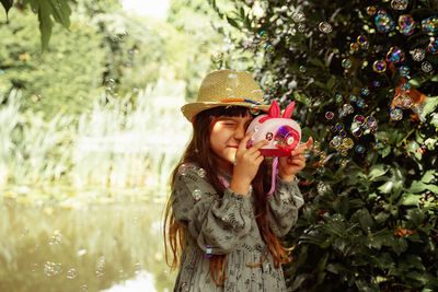 Portrait of young girl wearing hat standing and playing bubbles against tree