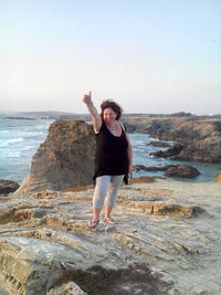 Portrait of woman showing thumbs up while standing on rock formation by sea against clear sky