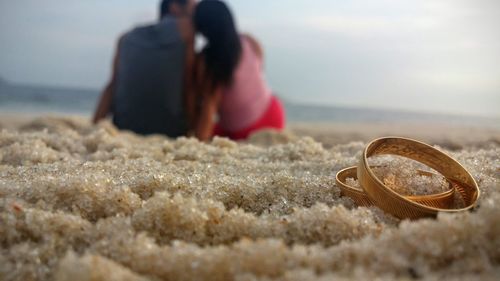 Pair of wedding rings on sand with couple sitting in background