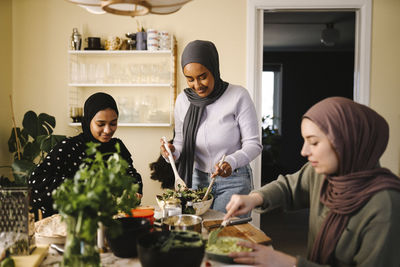 Smiling woman making salad while standing by friends preparing food at table in kitchen