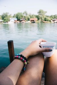 Midsection of woman holding cup while sitting by lake against sky