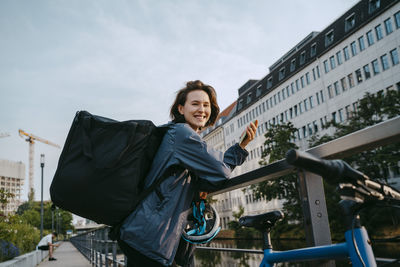 Side view portrait of smiling female delivery person holding smart phone while leaning on railing in city