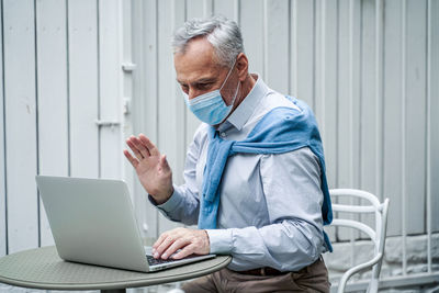 Smiling senior man wearing mask video calling while sitting at cafe