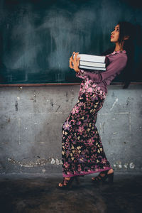 Side view of woman carrying books while walking against blackboard in classroom