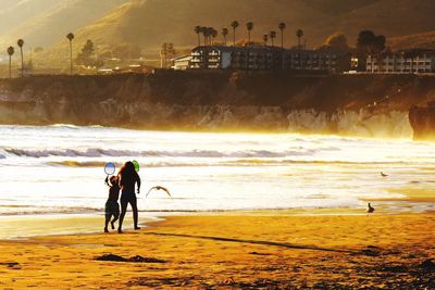 Rear view of woman with son running at beach against sky during sunset