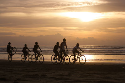 Silhouette people riding bicycles on beach against sky during sunset