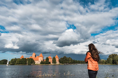 One woman standing on a lakeside with trakai castle in front, against cloudy sky