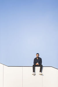 Man using phone while sitting on built structure against blue sky