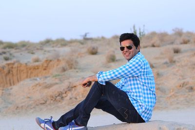 Portrait of young man sitting on retaining wall against clear sky