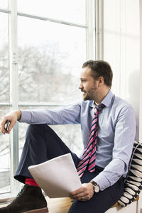 Mid adult businessman looking through window while sitting on ledge