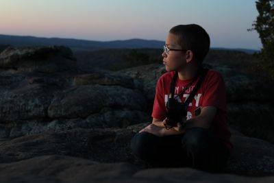 Boy sitting on rock against sky during sunset